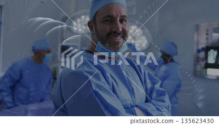 Standing lead surgeon in blue scrubs folding arms in operating room, with overhead lights, monitors 135623430