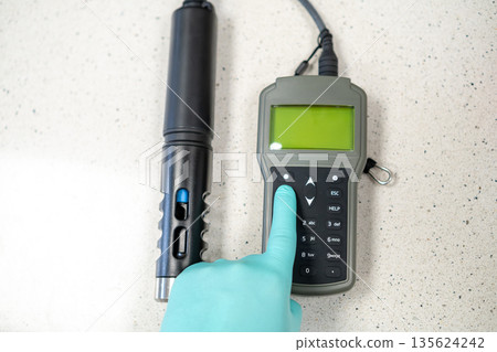 Female hands in protective gloves adjusting settings on a handheld pH meter. Female hands in protective gloves adjusting settings on a handheld pH meter. 135624242