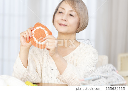 一位老婦人在客廳裡用羊毛線織一個腈綸材質的洗碗巾。 135624335