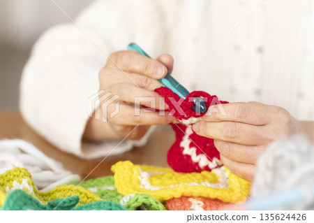 Out-of-focus hands of a senior woman knitting a woolen acrylic scrubber in the living room 135624426