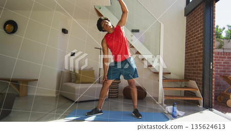 Performing standing side stretch man in red top on blue mat at home, with water bottle Performing standing side stretch man in red top on blue mat at home, with water bottle 135624613