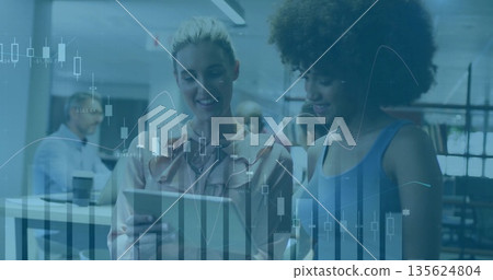 Two colleagues in blouse and sleeveless top reviewing tablet in office, with monitors, coffee cups 135624804