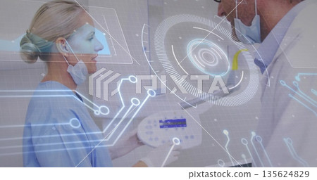 Female in scrubs holding control panel, lab-coat doctor viewing holograms in clinic, masks lowered 135624829
