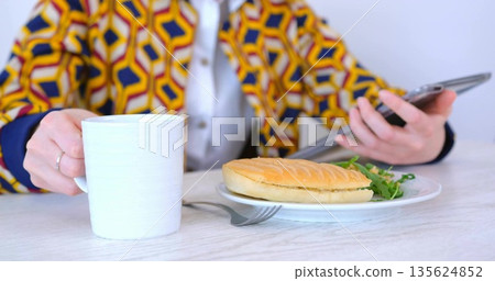 Holding white mug scrolling tablet wearing printed jacket at cafe table with panini fork and greens 135624852
