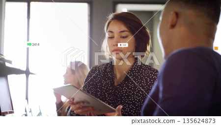 Talking woman in dark blouse holding tablet in open-plan office, grid-style windows and monitors 135624873