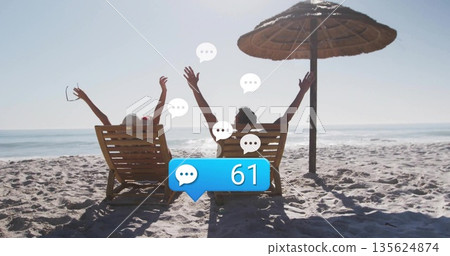 Raising arms couple celebrating on beach, with umbrella, sunglasses and chat notification bubbles 135624874
