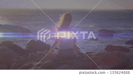Sitting lone woman facing calm ocean water at rocky shoreline during sunset, with lens flare Sitting lone woman facing calm ocean water at rocky shoreline during sunset, with lens flare 135624888
