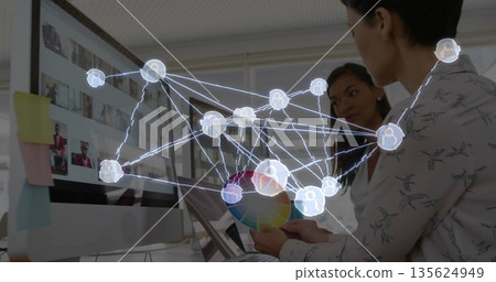 Two women in blouses holding color wheel and watching monitor at office desk, with network overlay 135624949