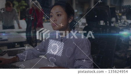 Typing mid adult Asian woman focusing on desktop monitor in open-plan office, with digital overlays 135624954