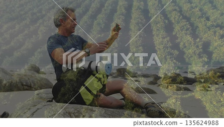 Mature hiker examining smartphone screen on large rock at coastal shore, with backpack and mat Mature hiker examining smartphone screen on large rock at coastal shore, with backpack and mat 135624988