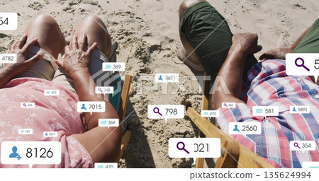 Sitting seniors wearing floral top and plaid shirt on deckchairs on sand with social media icons Sitting seniors wearing floral top and plaid shirt on deckchairs on sand with social media icons 135624994