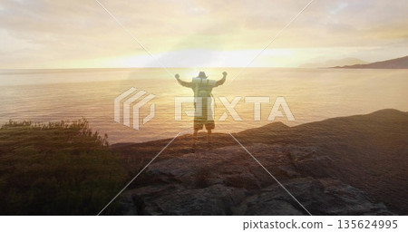 Raising arms hiker overlooking calm sea on headland at sunset, with backpack and wide-brimmed hat Raising arms hiker overlooking calm sea on headland at sunset, with backpack and wide-brimmed hat 135624995