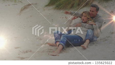 Couple in jackets wrapping arms around partner pointing right on coastal dune with dune grasses Couple in jackets wrapping arms around partner pointing right on coastal dune with dune grasses 135625022