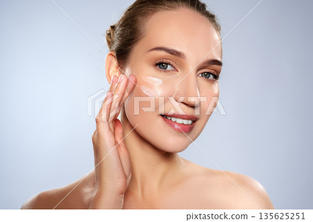 Woman applying skincare product on her face in a simple indoor setting Woman applying skincare product on her face in a simple indoor setting 135625251