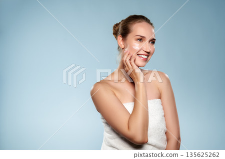 Woman smiling while applying skincare product in a simple setting with light blue background 135625262