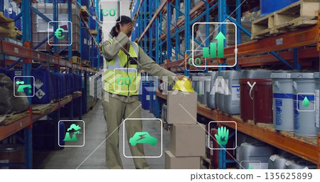 Surveying warehouse worker wearing safety vest holding hardhat shading eyes among drums, with icons 135625899