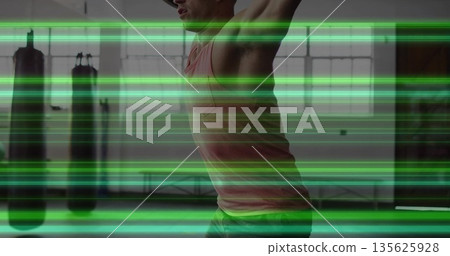 Stretching man in tank top raising arms overhead in gym, with punching bags, windows, padded floor 135625928