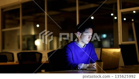 Working man wearing blue sweater typing on laptop in office at night, with paper coffee cup Working man wearing blue sweater typing on laptop in office at night, with paper coffee cup 135625992