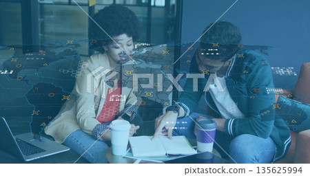 Reviewing documents, colleagues leaning over table in office lounge with laptop and map overlay 135625994