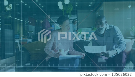 Two seated colleagues reviewing printed documents on sofa in office lounge, holding coffee cup 135626022