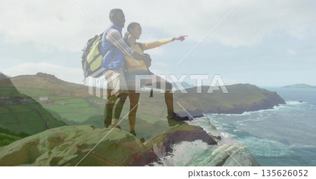 Leaning couple in yellow windbreaker embracing and pointing to sea from cliff, with hiking backpack 135626052