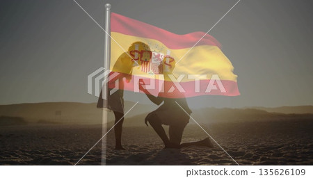 Proposing couple kneeling holding ring box on beach at sunset, with Spanish flag overlay 135626109