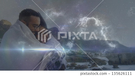 Embracing couple standing on rocky shoreline under starry sky, with large woven blanket Embracing couple standing on rocky shoreline under starry sky, with large woven blanket 135626111