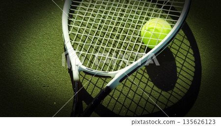 Lying diagonally white-framed tennis racket and yellow tennis ball casting shadows on green court 135626123