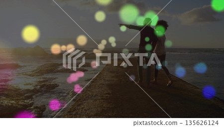 Pointing couple standing on stone pier at sunset, overlooking rocky shoreline with colored bokeh Pointing couple standing on stone pier at sunset, overlooking rocky shoreline with colored bokeh 135626124