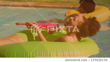 Woman in swimsuit aiming pink water gun on green tube in backyard pool, with yellow ring Woman in swimsuit aiming pink water gun on green tube in backyard pool, with yellow ring 135626130