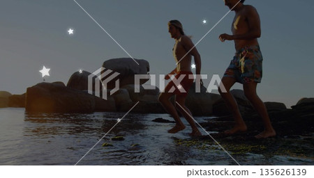 Stepping two friends in swim trunks exploring tide pools at water's edge, with boulders and seaweed Stepping two friends in swim trunks exploring tide pools at water's edge, with boulders and seaweed 135626139