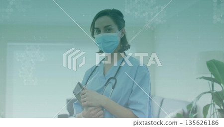 Standing nurse wearing light blue scrubs and mask in clinic, holding smartphone and stethoscope Standing nurse wearing light blue scrubs and mask in clinic, holding smartphone and stethoscope 135626186