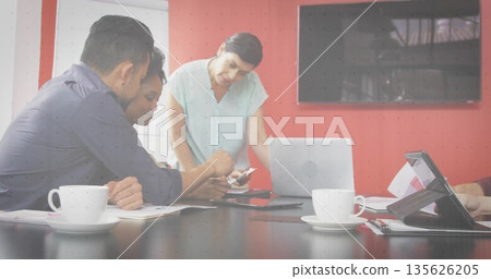 Collaborating professionals leaning over documents and devices at conference table, with coffee Collaborating professionals leaning over documents and devices at conference table, with coffee 135626205