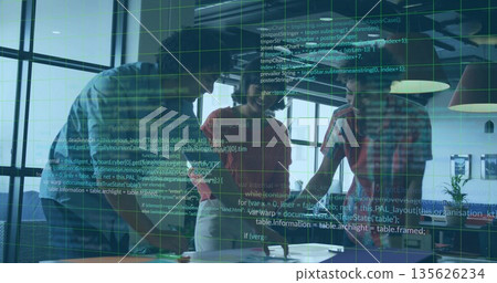 Collaborating professionals leaning over table in sunlit office, with laptops tablet documents 135626234