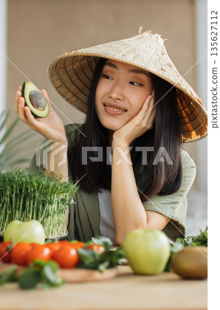A young woman wearing a traditional conical hat holds a halved avocado while looking thoughtfully to the side, surrounded by fresh produce 135627112