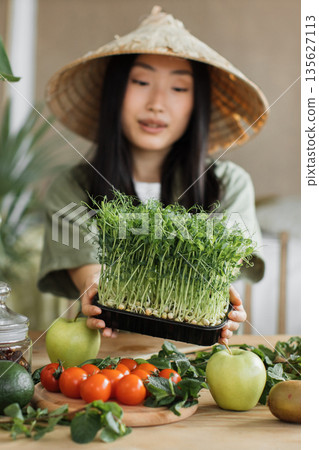 A young woman wearing a conical hat proudly presents a tray of freshly grown microgreens, surrounded by an assortment of healthy fruits and vegetables on a wooden table 135627113