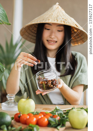 A young Asian woman wearing a traditional conical hat holds a jar of dates and a single date, surrounded by fresh produce on a wooden table A young Asian woman wearing a traditional conical hat holds a jar of dates and a single date, surrounded by fresh produce on a wooden table 135627117