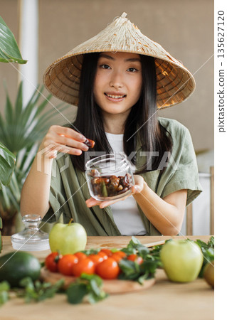 A young Asian woman wearing a traditional conical hat holds a jar of dates and prepares fresh produce for a healthy meal A young Asian woman wearing a traditional conical hat holds a jar of dates and prepares fresh produce for a healthy meal 135627120
