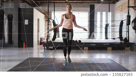 Jumping athlete wearing pink tank top skipping rope in boxing gym, with punching bags and gloves Jumping athlete wearing pink tank top skipping rope in boxing gym, with punching bags and gloves 135627165