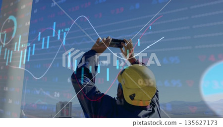 Field worker in coveralls and helmet raising smartphone at site, with financial chart overlay Field worker in coveralls and helmet raising smartphone at site, with financial chart overlay 135627173