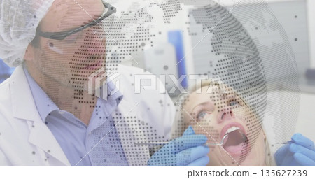 Examining male dentist leaning over female patient in dental clinic, with mirror and globe overlay 135627239