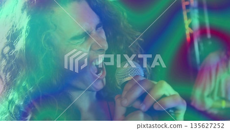 Singing curly-haired singer belting into microphone on club, with stage lights, truss and drum kit 135627252