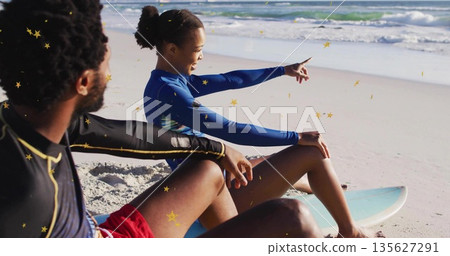 Sitting two surfers pointing toward ocean on pale sand at beach, with blue and red surfboards Sitting two surfers pointing toward ocean on pale sand at beach, with blue and red surfboards 135627291