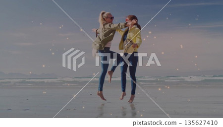 Leaping friends wearing olive and yellow jackets on beach near water's edge, with crossbody purses Leaping friends wearing olive and yellow jackets on beach near water's edge, with crossbody purses 135627410