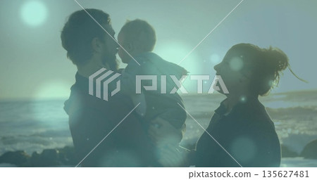 Cradling infant touching face, parents in dark tops posing at rocky beach sunset with ocean waves Cradling infant touching face, parents in dark tops posing at rocky beach sunset with ocean waves 135627481