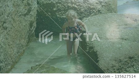 Exploring girl wearing star-print swimsuit peering at water on ledge in tide pool between boulders 135627514