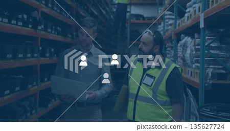 Three workers in safety gear retrieving stock on ladder in storage aisle, with laptop and documents 135627724