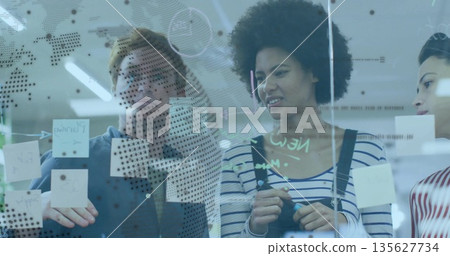 Collaborating colleagues placing sticky notes on glass wall in office, with dots and diagrams Collaborating colleagues placing sticky notes on glass wall in office, with dots and diagrams 135627734