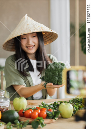 A young Asian woman wearing a conical hat holds a broccoli while filming herself on a smartphone, surrounded by fresh vegetables on a wooden table A young Asian woman wearing a conical hat holds a broccoli while filming herself on a smartphone, surrounded by fresh vegetables on a wooden table 135627761