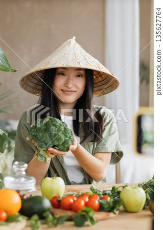 A young Asian woman wearing a conical hat holds a fresh broccoli head surrounded by various colorful vegetables on a wooden table A young Asian woman wearing a conical hat holds a fresh broccoli head surrounded by various colorful vegetables on a wooden table 135627764